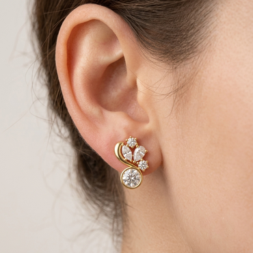 Model wearing an asymmetrical yellow gold diamond stud earring featuring a bezel-set round solitaire at the base with a sweeping gold ribbon and multi-shape diamond cluster above, shown close-up on the earlobe.