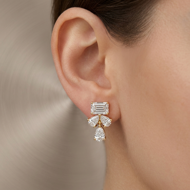 Model wearing a yellow gold emerald-cut diamond stud earring anchored by a three-stone cluster of pear-cut diamonds suspended below, shown close-up on the earlobe.
