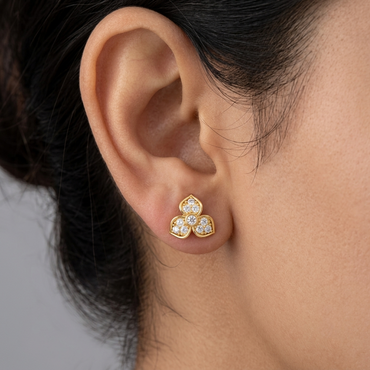 Model wearing a yellow gold trefoil floral stud earring with three pavé diamond-set petals centered around a bezel-set round diamond, shown close-up on the earlobe.