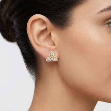Close-up of a model wearing a unique three-cluster yellow gold and diamond earring. The stud features a triple cluster of haloed marquise and pear-cut diamonds, arranged to form a trifoil or stylized fleur-de-lis. Model has dark hair pulled back and a neutral profile. White background.