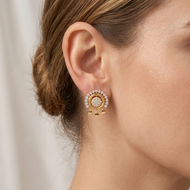 A gold and diamond earring with a detachable semi-circle jacket, worn on a model's ear. This ornate stud earring features pave-set diamonds and dangling gold charms. A model's face and shoulder are partially visible, with hair in a bun.